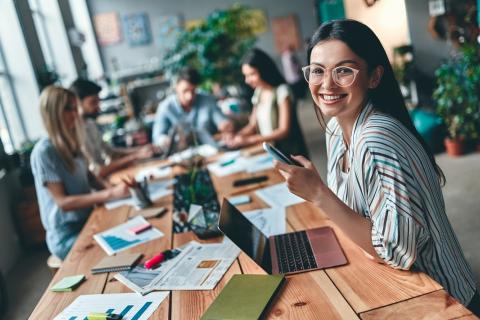 Jeune femme souriante, assise devant un ordinateur, avec un téléphone portable dans la main.