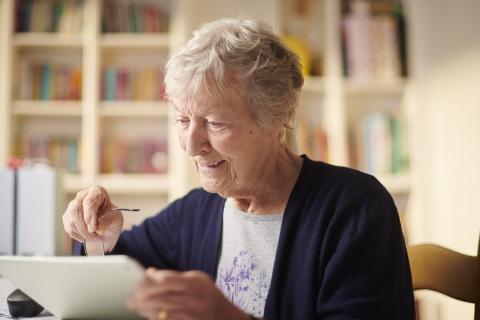 personne âgée assise à une table, en train de naviguer sur une tablette.