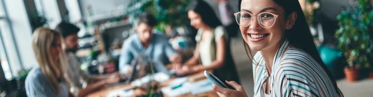 Jeune femme souriante, assise devant un ordinateur, avec un téléphone portable dans la main.