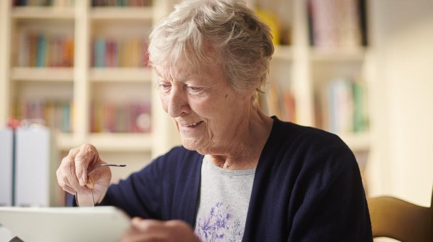 personne âgée assise à une table, en train de naviguer sur une tablette.